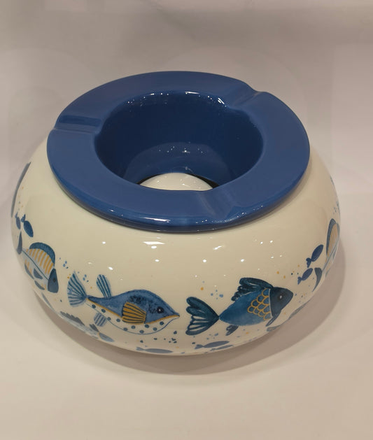 Posacenere antivento in ceramica con coperchio rimovibile blu lucido, decorato esternamente con un motivo artistico di pesciolini dipinti nelle tonalità dell'azzurro e del giallo. La struttura bombata color panna presenta tre scanalature per appoggiare le sigarette, rendendo questo accessorio per fumatori sia funzionale che perfetto come originale complemento d'arredo a tema marino.