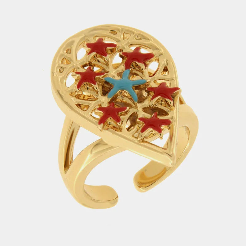 Anello regolabile con finitura dorata del marchio BySimon appartenente alla collezione Fiori di Lava caratterizzato da un importante castone a forma di goccia con lavorazione traforata a trama floreale. Il design è impreziosito da sette stelle marine tridimensionali smaltate, di cui sei di colore rosso vibrante disposte lungo il profilo e una centrale di colore azzurro a contrasto. La struttura dell'anello presenta una doppia fascia lucida che si unisce in un gambo aperto sul retro, ideale per regolare la m