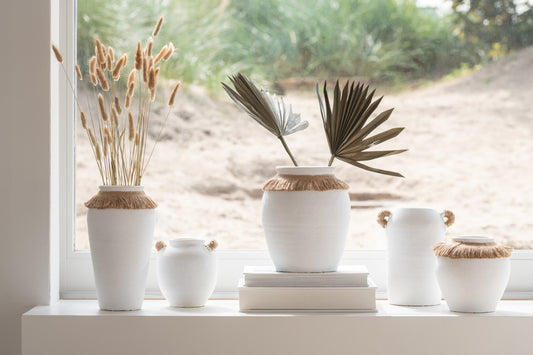 Vaso piccolo in ceramica della collezione Ibiza "Oceano Azul" di Jolipa, caratterizzato da una struttura bombata bianca con superficie dall'aspetto materico e naturale. Questo complemento d'arredo è decorato sulla parte superiore da un colletto in juta sfrangiata marrone, ideale per arricchire ambienti in stile boho chic o etnico.