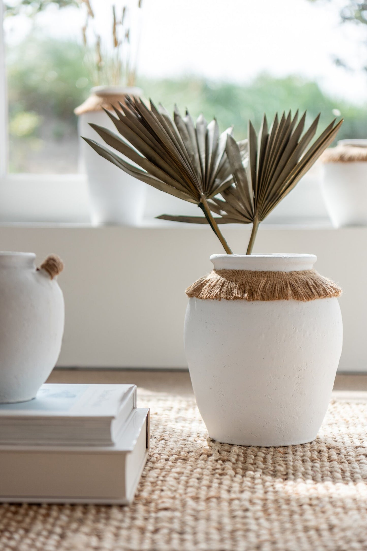 Vaso piccolo in ceramica della collezione Ibiza "Oceano Azul" di Jolipa, caratterizzato da una struttura bombata bianca con superficie dall'aspetto materico e naturale. Questo complemento d'arredo è decorato sulla parte superiore da un colletto in juta sfrangiata marrone, ideale per arricchire ambienti in stile boho chic o etnico.