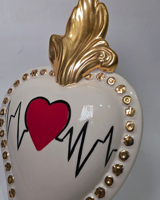 Scultura decorativa a forma di Cuore Sacro in ceramica bianca lucida, caratterizzata da un cuore rosso centrale sovrapposto a un disegno grafico nero che richiama il battito cardiaco. L'oggetto è impreziosito da una fiamma superiore dorata e una cornice perlinata con borchie oro, rendendolo un complemento d'arredo dal design moderno e originale.