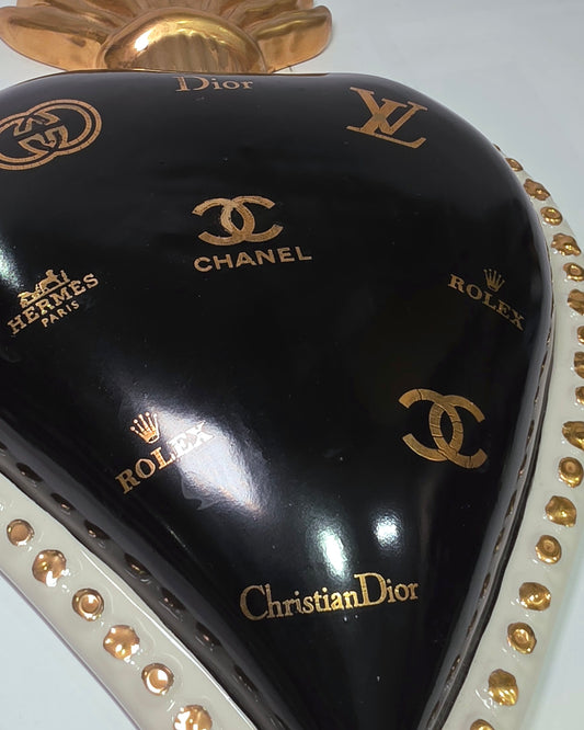 Scultura da parete a forma di Cuore Sacro grande in ceramica nera lucida, impreziosita da numerosi loghi fashion dorati e sormontata da una fiamma in oro finemente scolpita. Questo complemento d'arredo pop art presenta una cornice bianca perlinata con borchie oro, perfetto per decorare con lusso e originalità gli interni di una casa moderna.