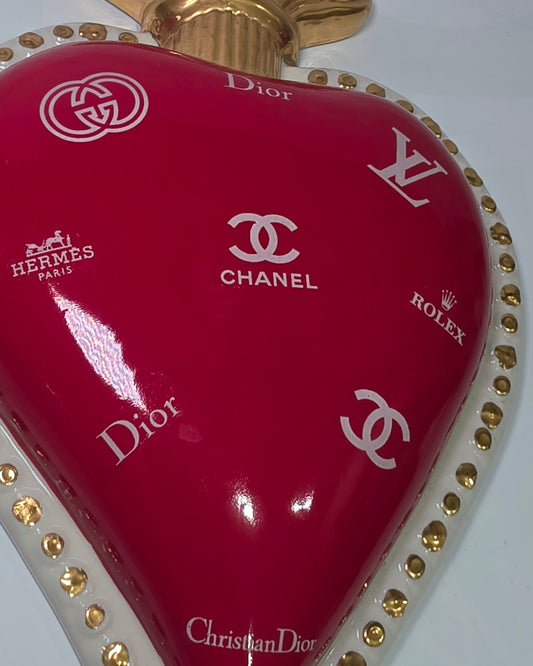 Scultura da parete a forma di Cuore Sacro grande in ceramica rossa lucida, decorata con diversi loghi fashion bianchi e sormontata da una fiamma dorata a rilievo. L'oggetto è rifinito con una cornice perlinata bianca e borchie oro, rappresentando un complemento d'arredo pop art dal design audace ideale per ambienti moderni.