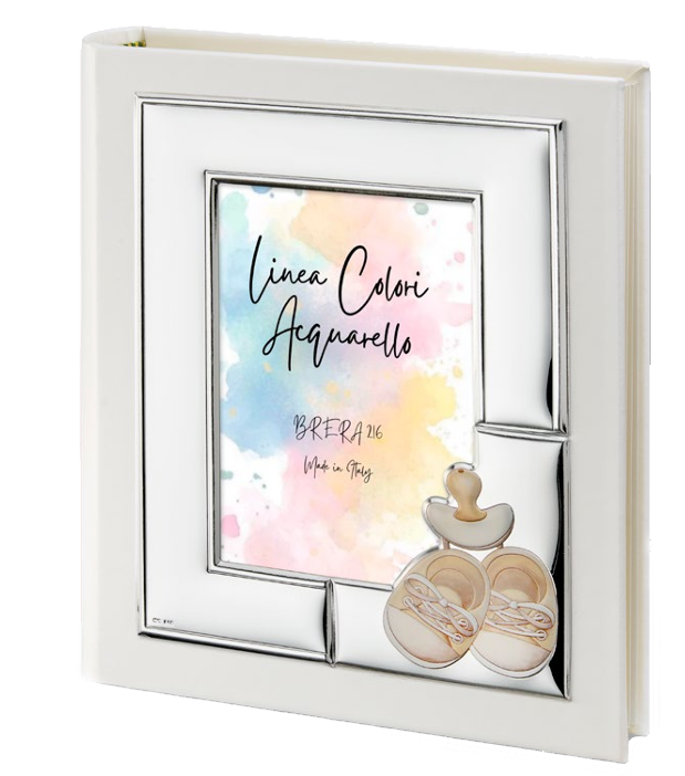 Album portafoto in ecopelle bianca con cornice frontale in argento bilaminato. La cornice è decorata nell'angolo in basso a destra con applicazioni in rilievo smaltate che raffigurano un paio di scarpette da neonato e un ciuccio nei toni del beige e panna. All'interno della cornice è visibile un inserto color acquerello con la scritta "Linea Colori Acquarello - Brera 216 - Made in Italy".