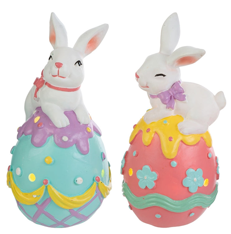Coppia di statuine pasquali in resina con LED. Ogni soggetto raffigura un coniglietto bianco disteso sopra un grande uovo decorato con colori vivaci, fiori e motivi a rilievo. Le uova presentano piccoli fori circolari da cui filtra la luce interna. Una versione ha l'uovo azzurro e viola, l'altra rosa e giallo.