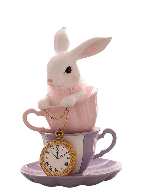 Statuina decorativa in resina con un coniglietto bianco seduto all'interno di una tazzina rosa a pois bianchi, impilata sopra una tazzina lilla a strisce. Il coniglio indossa un collarino di tulle rosa e tiene una catenella dorata collegata a un grande orologio da taschino appoggiato alla base. La composizione poggia su un piattino lilla sagomato.