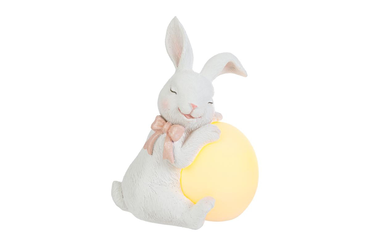 Statuina pasquale luminosa del marchio Le Stelle (codice 12206) raffigurante un coniglietto bianco seduto che abbraccia un grande uovo a LED giallo. Il coniglio ha gli occhi chiusi in un'espressione serena e indossa un fiocco rosa al collo. Decorazione da tavolo in resina con luce soffusa ideale per l'arredo casa.