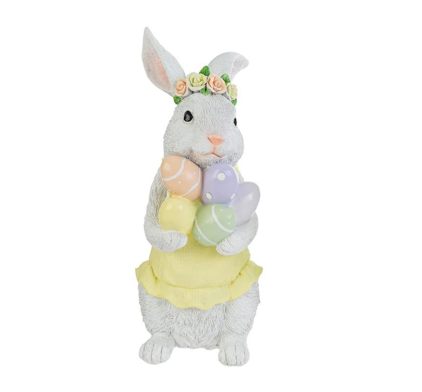 Statuina decorativa in resina del marchio Le Stelle (codice 12242) raffigurante una coniglietta bianca con una corona di roselline pastello sulla testa. La coniglietta indossa un vestitino giallo con balze e stringe tra le zampe un gruppo di uova di Pasqua colorate e decorate. Oggetto d'arredo primaverile con texture effetto pelo realistica.