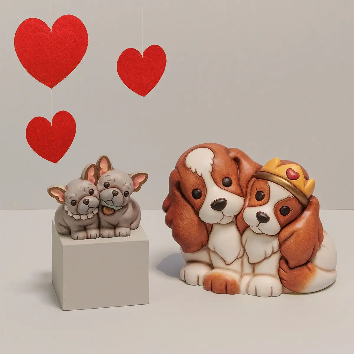 Composizione con la coppia di Cavalier King accanto a una statuina più piccola di Bulldog Francesi grigi su un piedistallo. Sullo sfondo pendono tre cuori rossi in feltro.