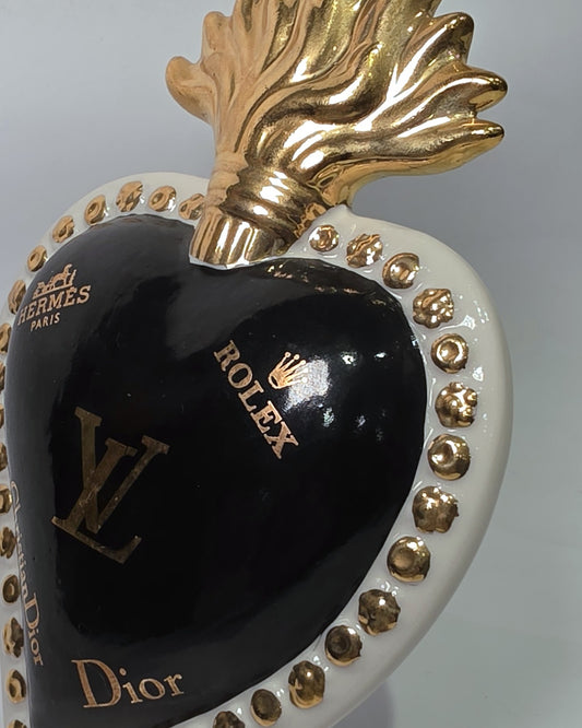 Scultura in ceramica a forma di Cuore Sacro firmata Sbordone, caratterizzata da un corpo nero lucido decorato con loghi fashion dorati e una fiamma superiore in oro. L'opera è rifinita da una cornice bianca perlinata con borchie dorate, rappresentando un complemento d'arredo pop art esclusivo e moderno per interni di design.