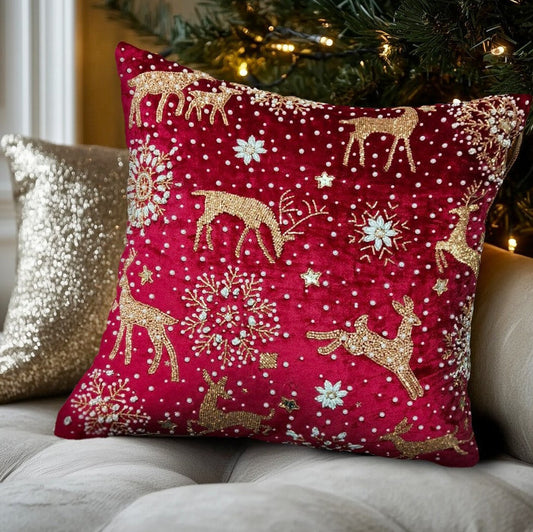 Inquadratura ravvicinata di un cuscino natalizio rosso ricamato con renne e fiocchi di neve, adagiato su un divano grigio accanto a un cuscino glitterato dorato. Sullo sfondo, luci di Natale soffuse.