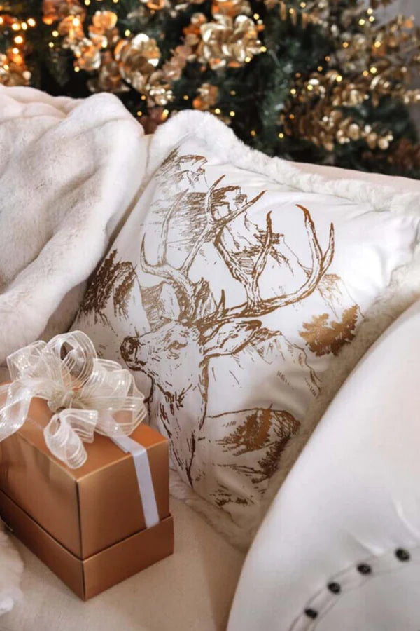 Cuscino natalizio bianco con stampa oro di un cervo e bordo in morbida pelliccia, appoggiato su un divano accanto a un pacchetto regalo dorato e un albero di Natale illuminato.