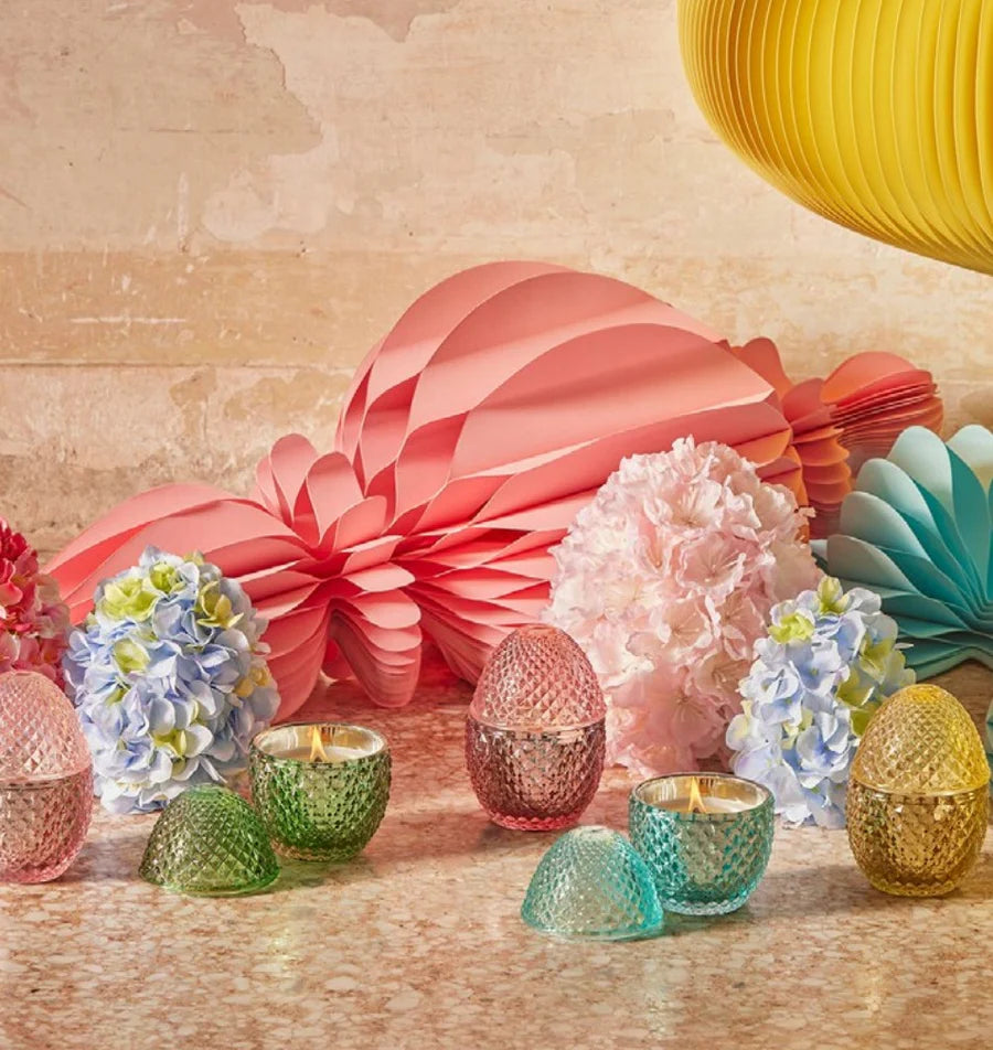 Immagine d'ambientazione per la collezione di candele profumate "L'Ove" firmata EDG Enzo de Gasperi, con eleganti contenitori in vetro colorato lavorato a rilievo a forma di uovo disposti tra decorazioni floreali e festoni di carta colorati. Questa linea di candele decorative di lusso, mostrata sia aperte e accese che con coperchio coordinato, è ideale per profumare gli interni con eleganza e creare un'atmosfera allegra, ricercata e primaverile.