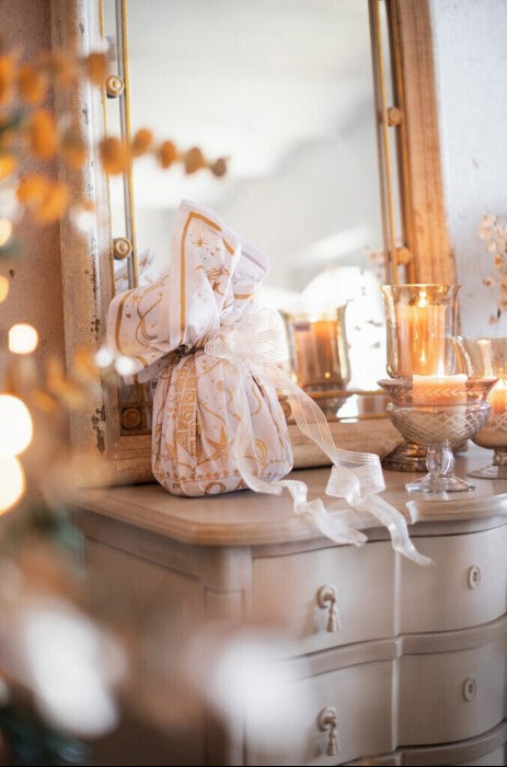 Panettone confezionato con foulard bianco e oro su un mobile antico con specchio. Atmosfera natalizia calda con candele accese e decorazioni luminose sfocate in primo piano.