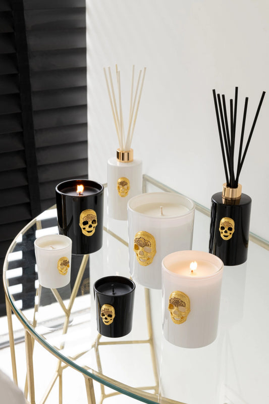 mmagine d'ambientazione della linea di profumatori e candele "Cranio" Nuit Noire firmata Jolipa, che mostra una raffinata selezione di prodotti disposti su un tavolino di design in vetro e metallo dorato. La collezione, caratterizzata dall'inconfondibile dettaglio a rilievo a forma di teschio dorato, accosta elementi bianchi e neri lucidi, tra cui diffusori di fragranza con bastoncini coordinati e candele profumate di varie dimensioni.