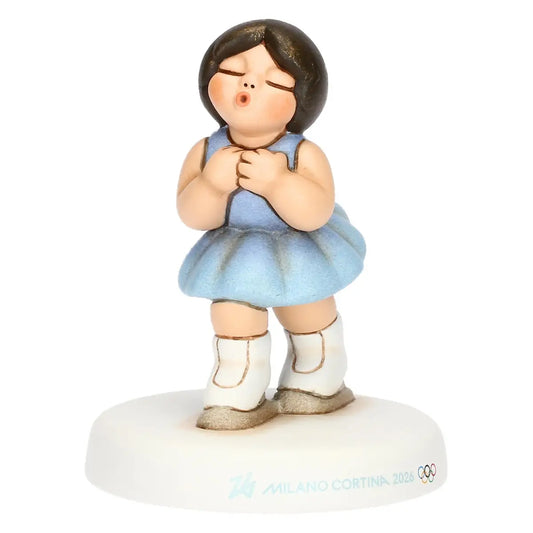 Vista a tre quarti della pattinatrice in ceramica Milano Cortina 2026. L'angolazione mette in risalto la posa aggraziata, i dettagli dell'abito azzurro e i loghi sulla base.