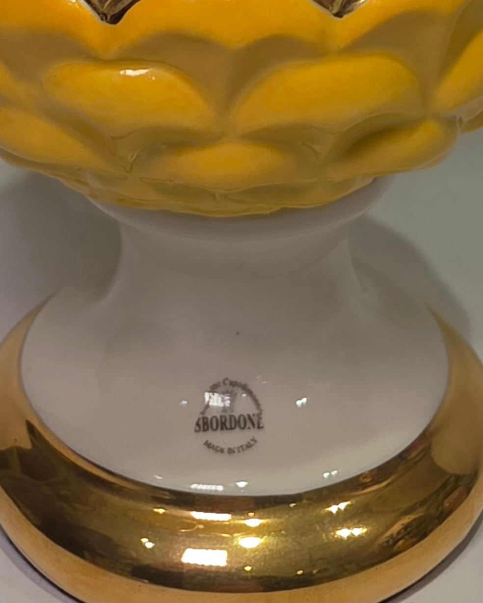 Pigna portafortuna in ceramica bicolore gialla e bianca lucida, impreziosita da una raffinata corona di squame dorate che ne definisce il profilo centrale. Questo elegante oggetto ornamentale artigianale poggia su una base circolare con finiture in oro, rappresentando un complemento d'arredo perfetto per decorazioni d'interni in stile chic e contemporaneo.