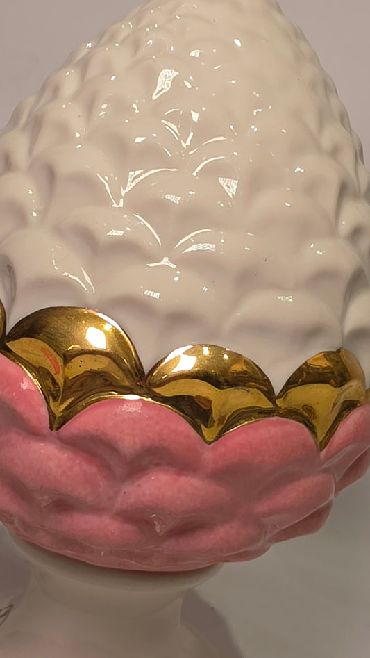 Pigna portafortuna in ceramica bicolore rosa e bianca lucida, impreziosita da una raffinata corona di squame dorate che ne definisce il profilo centrale. Questo elegante oggetto ornamentale artigianale poggia su una base circolare con finiture in oro, rappresentando un complemento d'arredo perfetto per decorazioni d'interni in stile chic e contemporaneo.