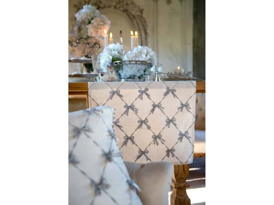 Dettaglio di un runner Blanc Mariclò con fiocchi ricamati grigio argento. Il tessuto cade lateralmente da un tavolo in legno, in un contesto chic con candele accese e ortensie bianche sfocate.