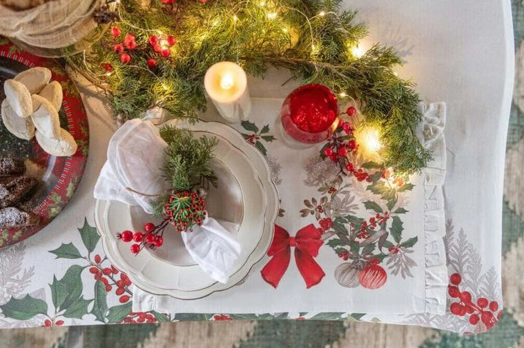Ambientazione natalizia con runner Sinfonia di Natale, piatti bianchi, tovagliolo con decoro di pino e bacche, candele e ghirlanda luminosa. Elegante mise en place festiva.