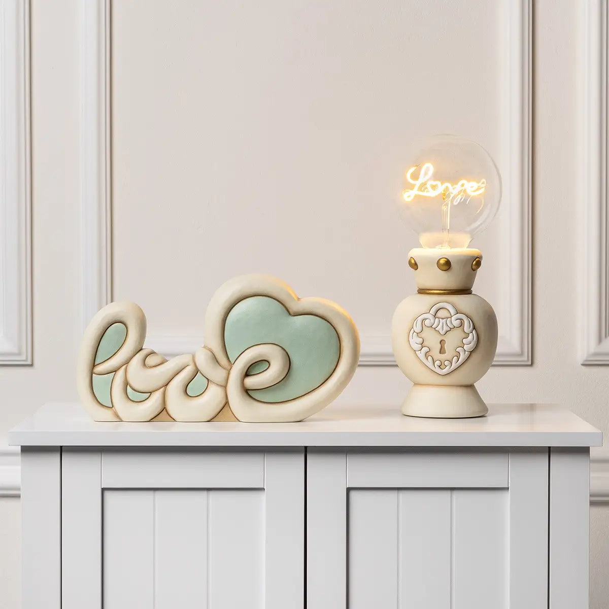 La decorazione bicolore (avorio e verde acqua) è posizionata su un mobile bianco accanto a una lampada in ceramica dotata di una lampadina a bulbo trasparente che proietta la parola "Love" come filamento luminoso.