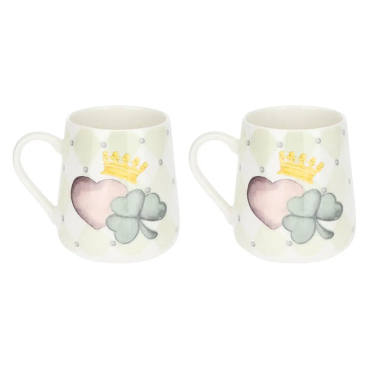 L'immagine mostra due tazze mug in ceramica identiche, posizionate l'una accanto all'altra su uno sfondo bianco neutro. Entrambe le tazze presentano un design a rombi in delicate tonalità di crema e verde chiaro, punteggiato da piccoli pois grigi. La decorazione principale, centrata sul corpo di ogni tazza, raffigura un cuore rosa sfumato e un quadrifoglio verde salvia in stile acquerello, sormontati da una corona gialla stilizzata.
