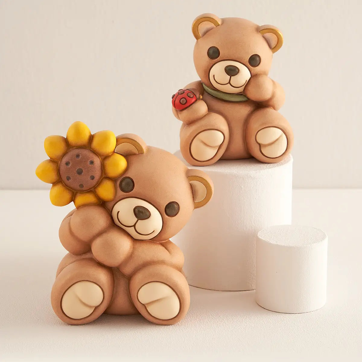 Due Teddy THUN in ceramica su basi cilindriche bianche: in primo piano il Teddy con girasole grande, sullo sfondo il Teddy con coccinella. Composizione decorativa per interni.