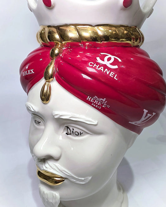 Coppia di Teste di Moro in ceramica bianca lucida con turbanti rosso ciliegia decorati da loghi fashion bianchi, completate da corone regali e labbra con finitura in oro zecchino. Queste sculture ornamentali di design pop art fondono la tradizione ceramica siciliana con l'estetica del lusso moderno, rappresentando un complemento d'arredo audace ed esclusivo per interni ricercati.