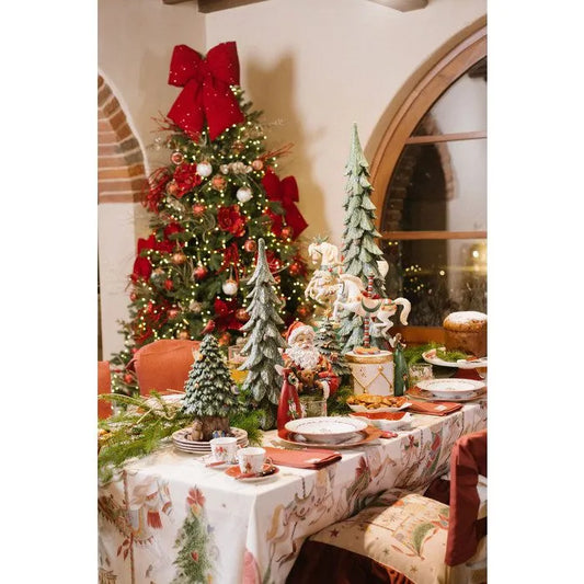 Elegante mise en place festiva con la tovaglia in cotone Nuvole di Stoffa "La Giostra di Amelie". La tavola è imbandita con stoviglie coordinate e decorata con alberi di Natale in miniatura e statuine, davanti a un grande albero di Natale addobbato con luci e un fiocco rosso.