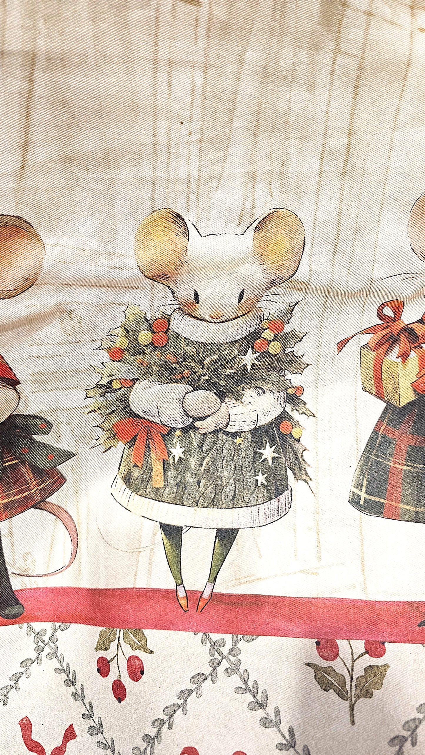 Primo piano della stampa su tessuto Blanc Mariclo: un tenero topino bianco indossa un maglione verde a trecce e tiene tra le zampe una ghirlanda di agrifoglio. Texture del tessuto visibile.