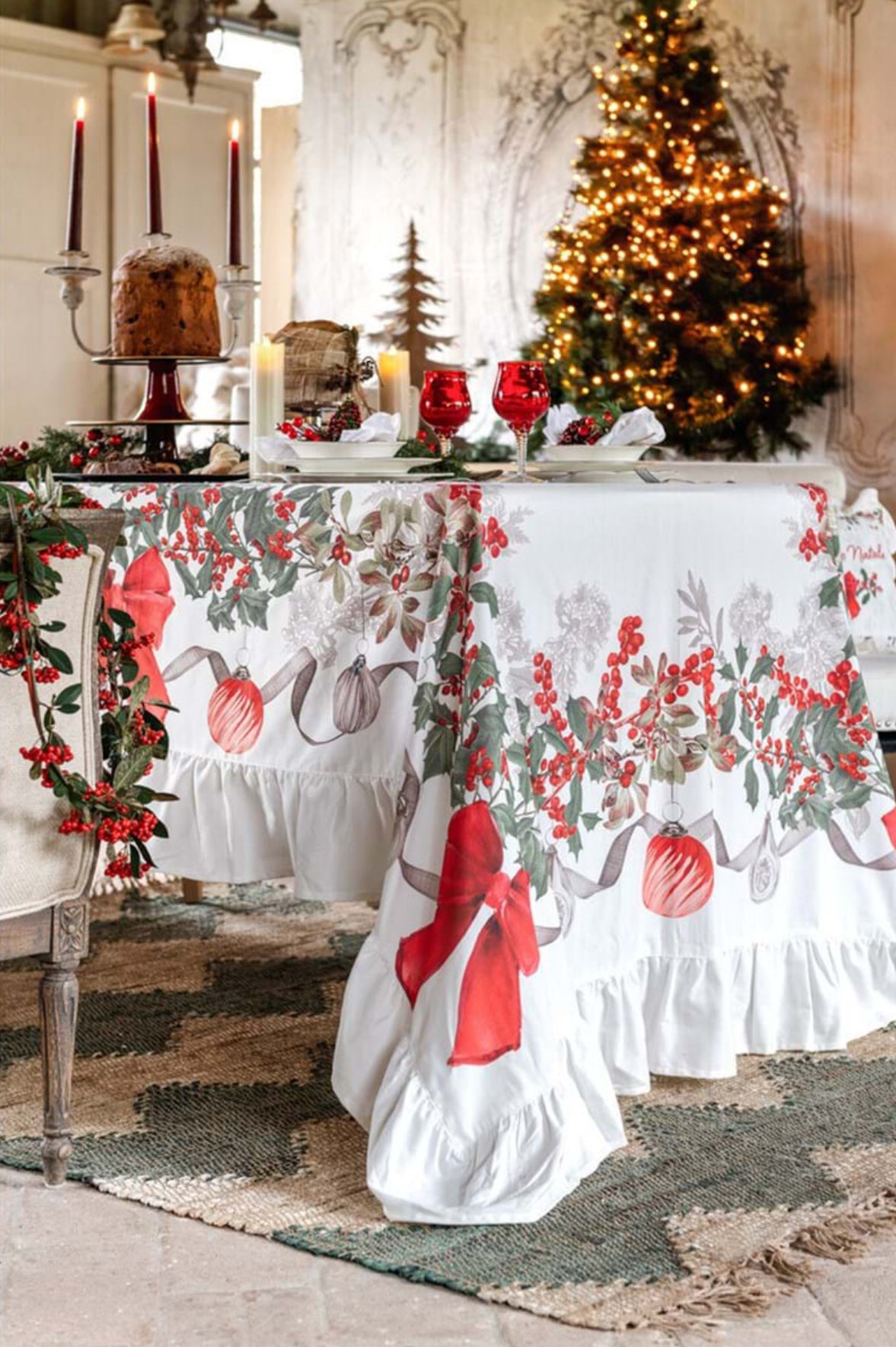 Tavola imbandita con tovaglia a gala Sinfonia Natalizia. Sul piano, calici rossi, candele e panettone. Sullo sfondo un albero di Natale illuminato e pareti decorate creano un'atmosfera calda ed elegante.