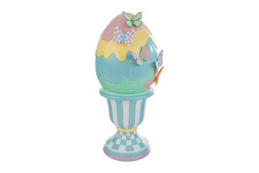 Decorazione pasquale a forma di uovo gigante su piedistallo a marchio Le Stelle. L'uovo presenta strati color pastello (rosa, giallo, azzurro e verde) con bordo a onde e farfalle decorative applicate. Il piedistallo è decorato con strisce bianche e azzurre e una base a scacchi celesti.