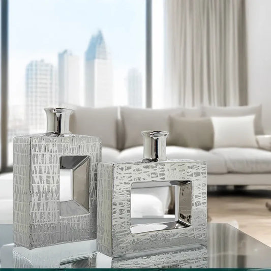 Vaso a bottiglia rettangolare orizzontale firmato Camacho, caratterizzato da un'elegante finitura ceramica color argento lucido con una texture a rilievo finemente lavorata e un originale design geometrico irregolare con foro centrale vuoto. Questo moderno complemento d'arredo è provvisto di un collo stretto superiore ideale per ospitare singoli steli floreali, perfetto come sofisticato oggetto ornamentale per impreziosire mobili, consolle o tavoli in contesti contemporanei.