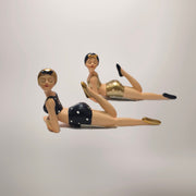 Coppia di statuine decorative assortite raffiguranti nuotatrici sdraiate a pancia in giù del marchio Efya, ideali come soprammobili estivi dal design retrò. Le due bagnanti con occhi chiusi e stile pin-up vintage indossano costumi da bagno a due pezzi abbinati a graziose cuffie, una in versione nera a pois bianchi e l'altra con finitura dorata lucida.