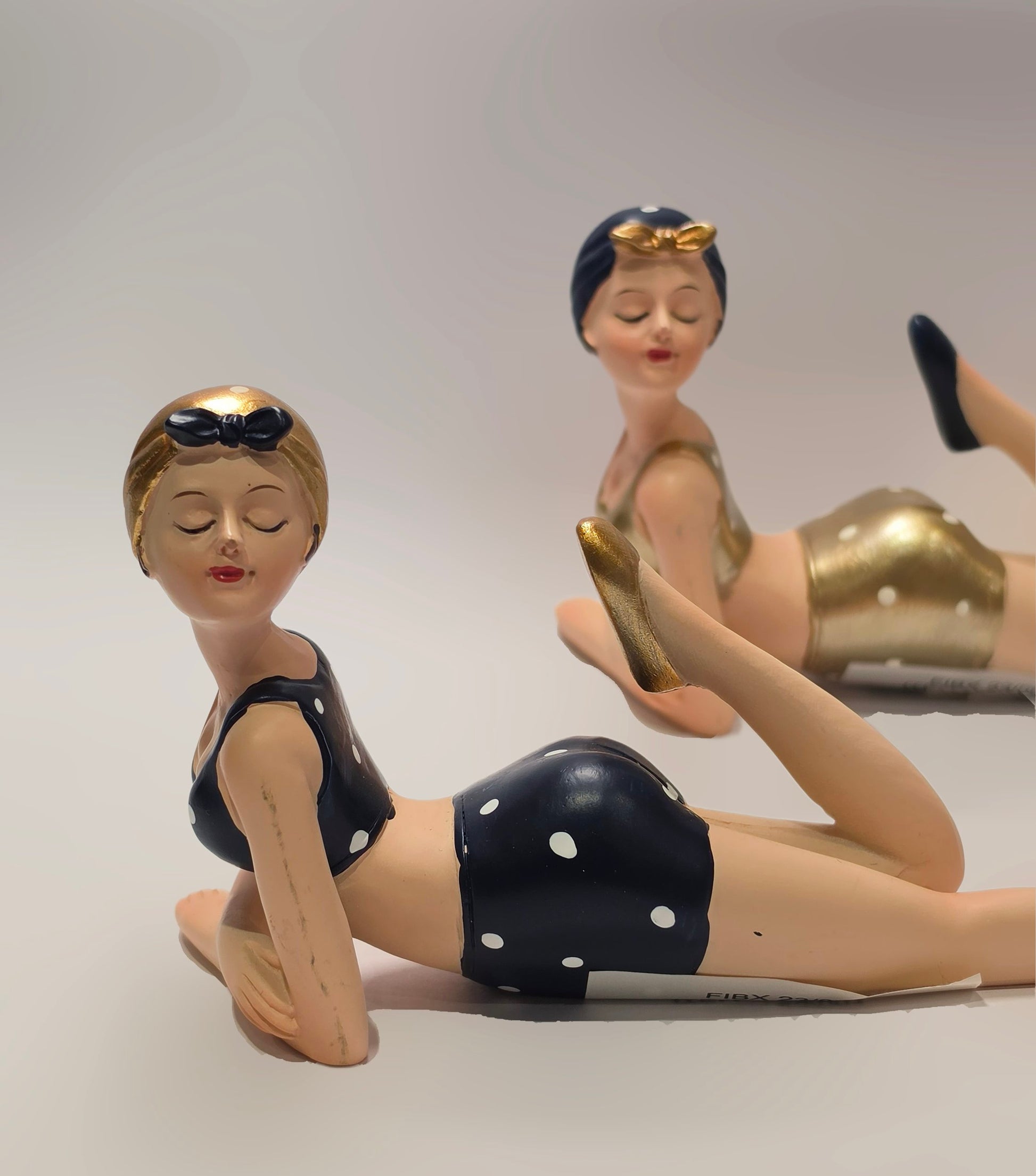 Coppia di statuine decorative assortite raffiguranti nuotatrici sdraiate a pancia in giù del marchio Efya, ideali come soprammobili estivi dal design retrò. Le due bagnanti con occhi chiusi e stile pin-up vintage indossano costumi da bagno a due pezzi abbinati a graziose cuffie, una in versione nera a pois bianchi e l'altra con finitura dorata lucida.