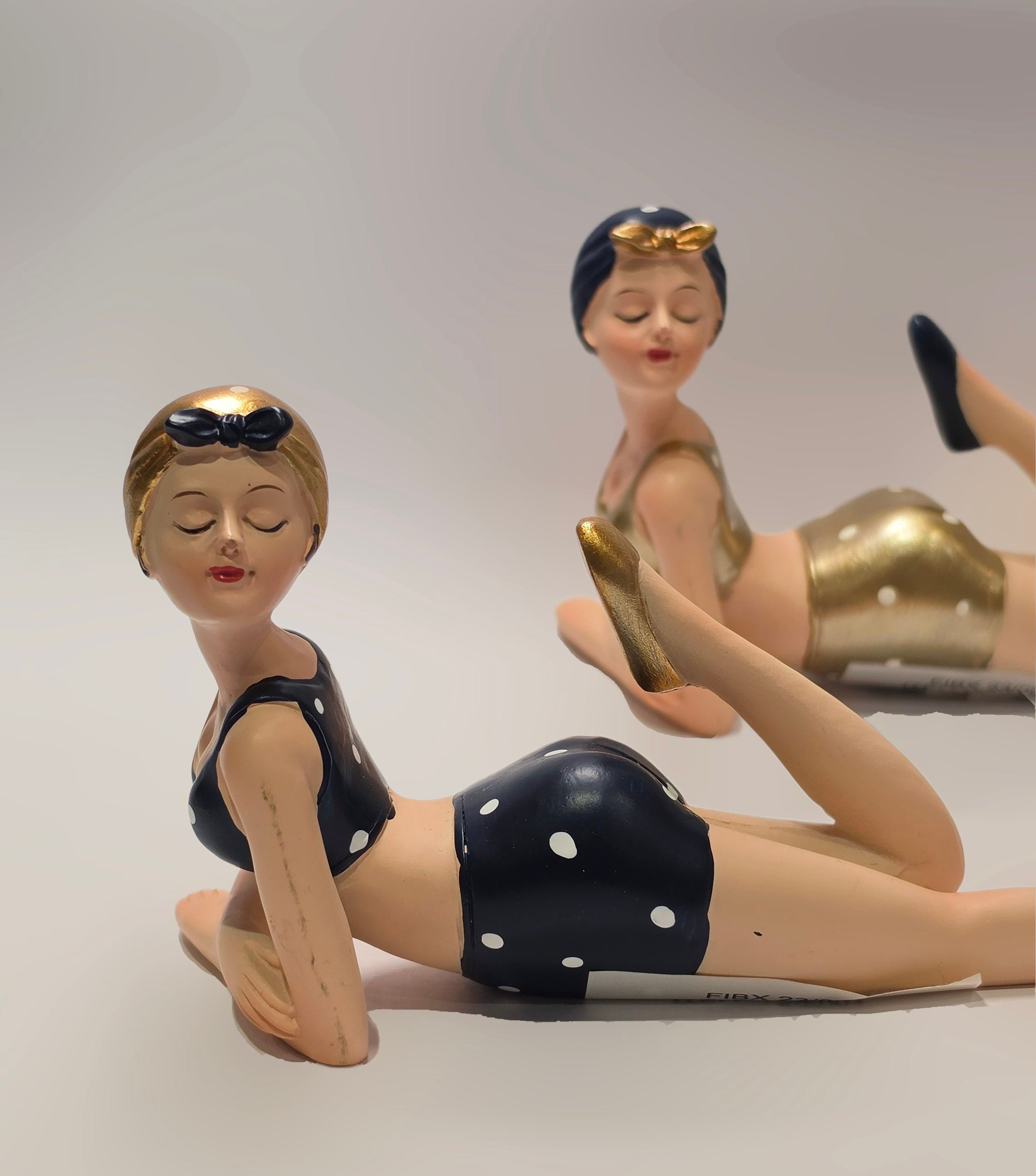Coppia di statuine decorative assortite raffiguranti nuotatrici sdraiate a pancia in giù del marchio Efya, ideali come soprammobili estivi dal design retrò. Le due bagnanti con occhi chiusi e stile pin-up vintage indossano costumi da bagno a due pezzi abbinati a graziose cuffie, una in versione nera a pois bianchi e l'altra con finitura dorata lucida.