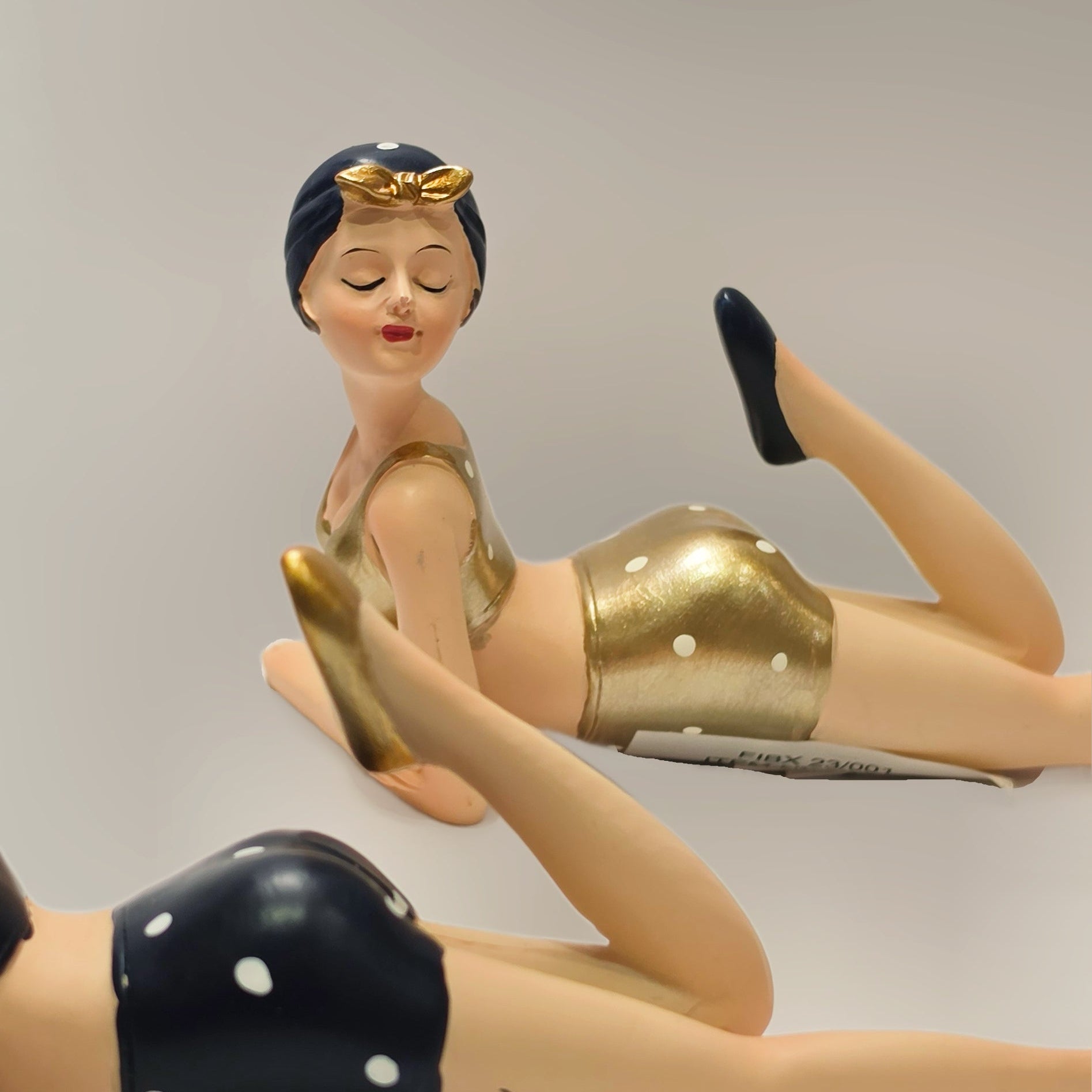 Coppia di statuine decorative assortite raffiguranti nuotatrici sdraiate a pancia in giù del marchio Efya, ideali come soprammobili estivi dal design retrò. Le due bagnanti con occhi chiusi e stile pin-up vintage indossano costumi da bagno a due pezzi abbinati a graziose cuffie, una in versione nera a pois bianchi e l'altra con finitura dorata lucida.