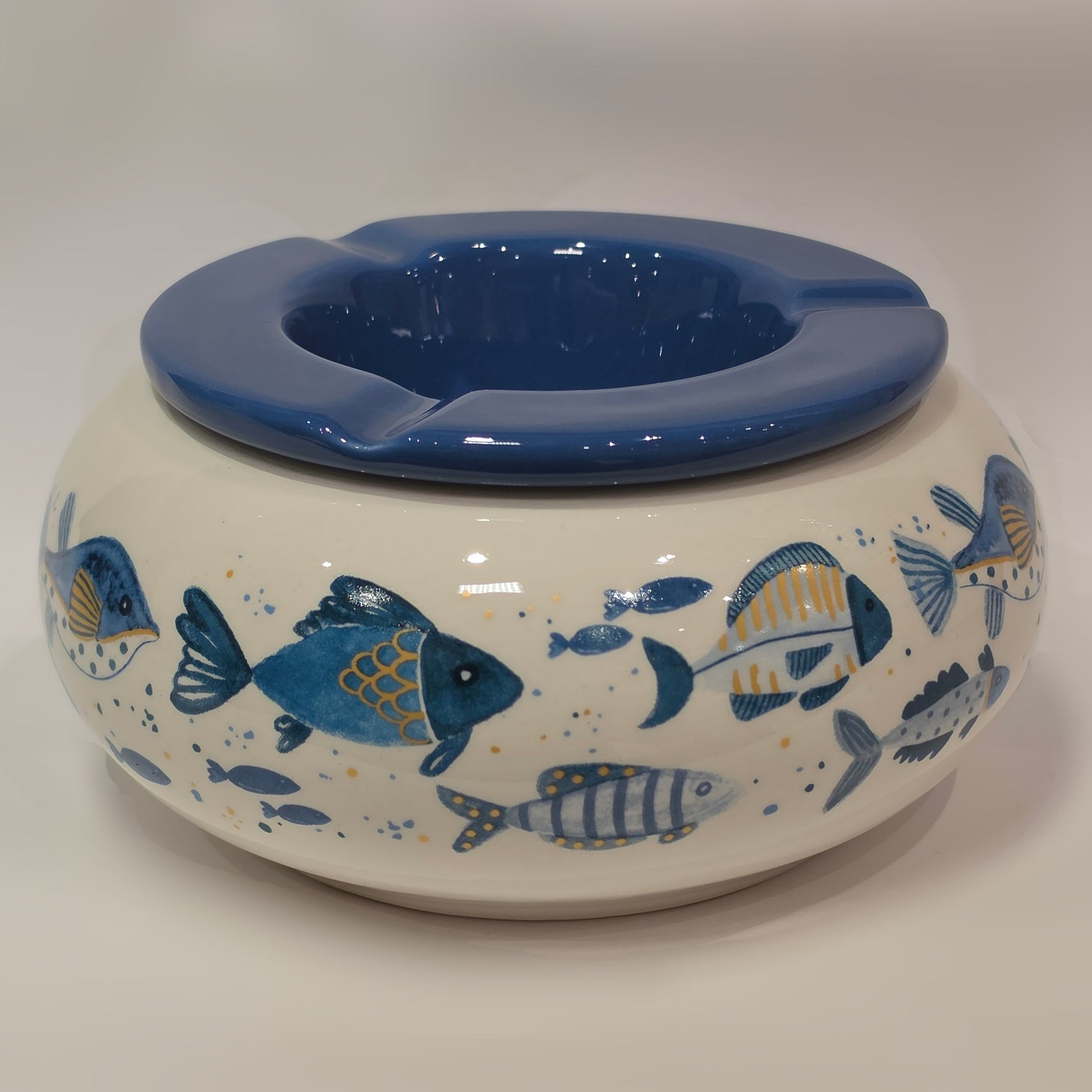 Posacenere antivento in ceramica con coperchio rimovibile blu lucido, decorato esternamente con un motivo artistico di pesciolini dipinti nelle tonalità dell'azzurro e del giallo. La struttura bombata color panna presenta tre scanalature per appoggiare le sigarette, rendendo questo accessorio per fumatori sia funzionale che perfetto come originale complemento d'arredo a tema marino.
