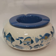 Posacenere antivento in ceramica con coperchio rimovibile blu lucido, decorato esternamente con un motivo artistico di pesciolini dipinti nelle tonalità dell'azzurro e del giallo. La struttura bombata color panna presenta tre scanalature per appoggiare le sigarette, rendendo questo accessorio per fumatori sia funzionale che perfetto come originale complemento d'arredo a tema marino.