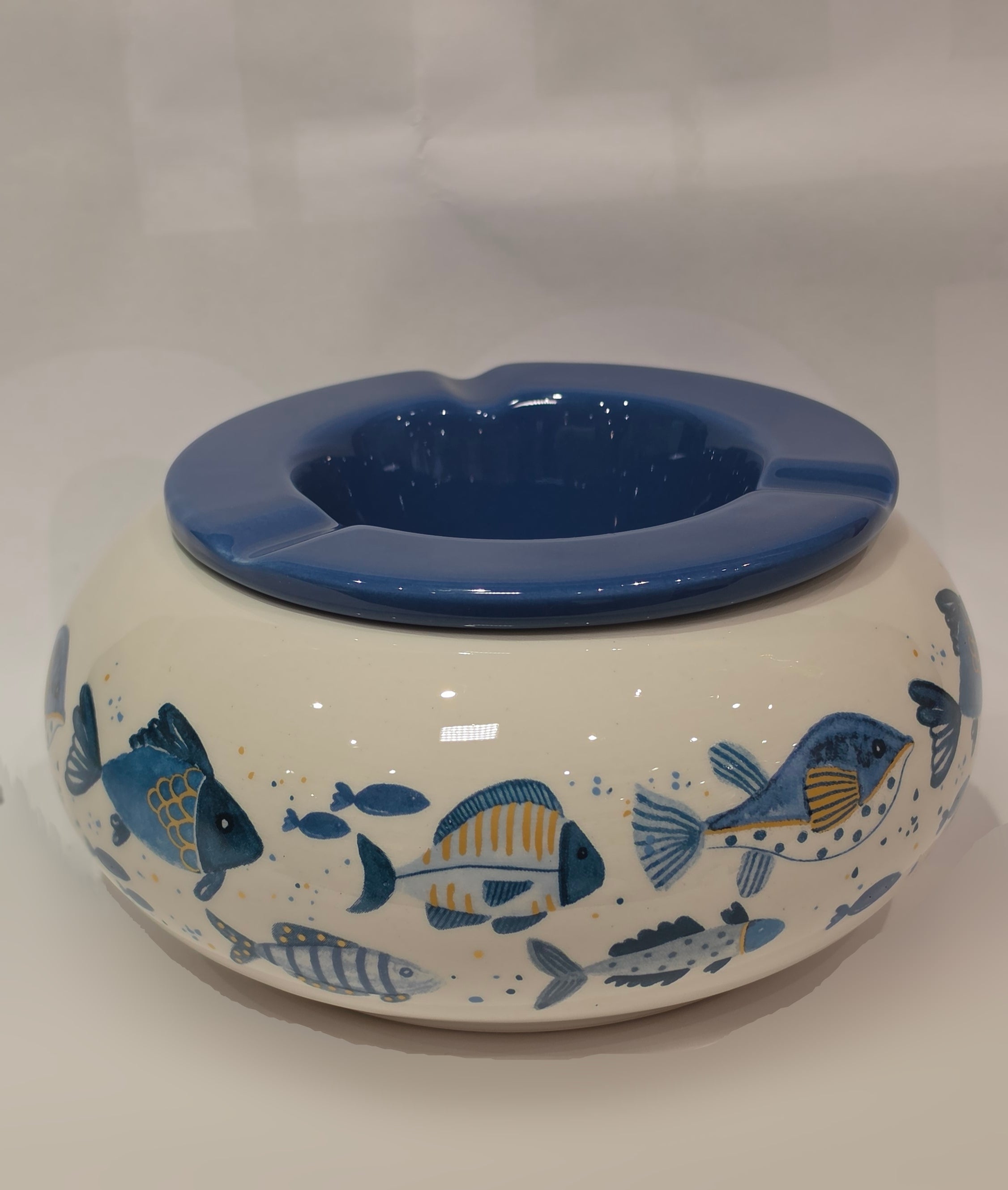Posacenere antivento in ceramica con coperchio rimovibile blu lucido, decorato esternamente con un motivo artistico di pesciolini dipinti nelle tonalità dell'azzurro e del giallo. La struttura bombata color panna presenta tre scanalature per appoggiare le sigarette, rendendo questo accessorio per fumatori sia funzionale che perfetto come originale complemento d'arredo a tema marino.