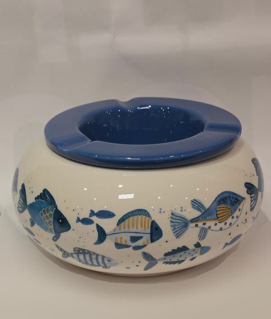 Posacenere antivento in ceramica con coperchio rimovibile blu lucido, decorato esternamente con un motivo artistico di pesciolini dipinti nelle tonalità dell'azzurro e del giallo. La struttura bombata color panna presenta tre scanalature per appoggiare le sigarette, rendendo questo accessorio per fumatori sia funzionale che perfetto come originale complemento d'arredo a tema marino.