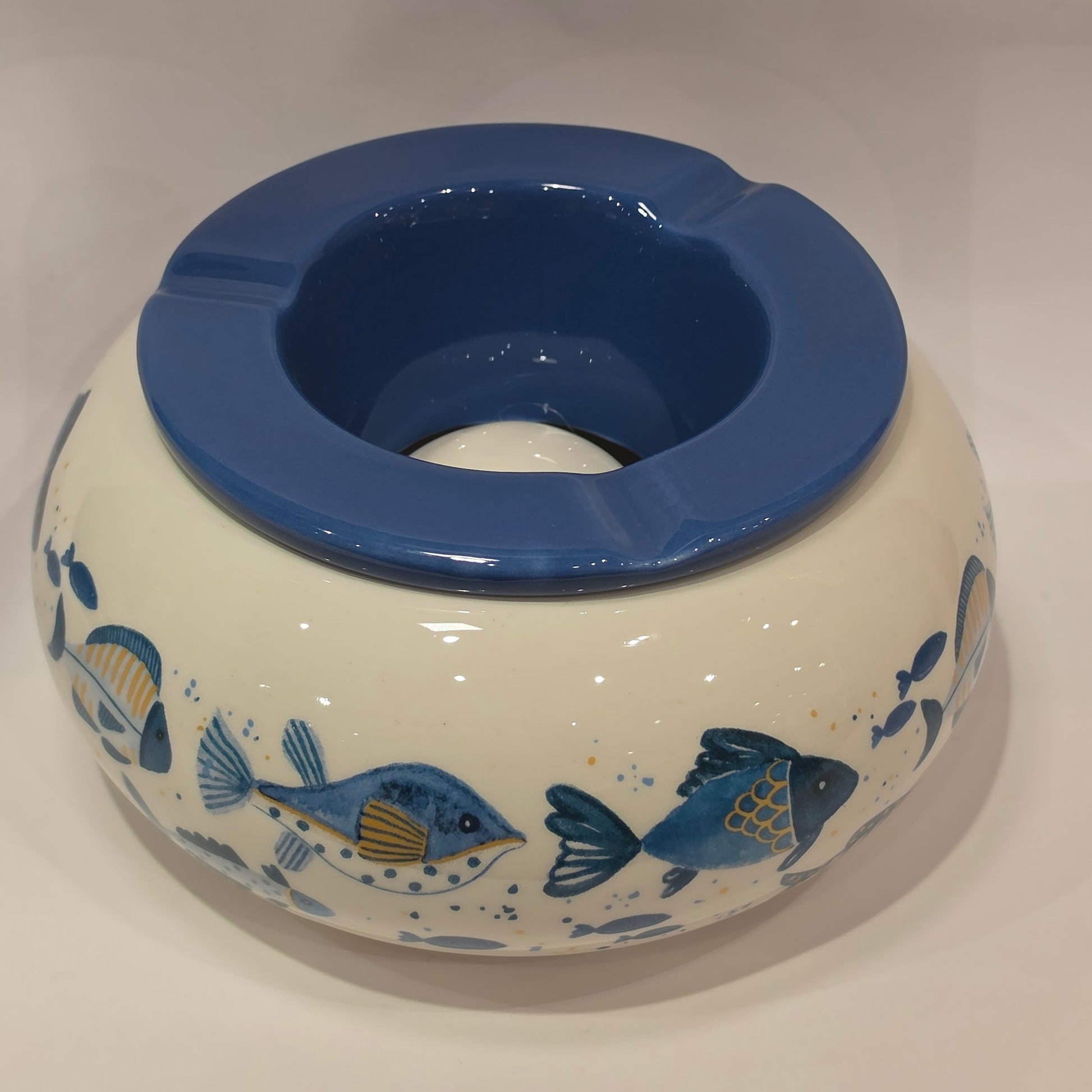 Posacenere antivento in ceramica con coperchio rimovibile blu lucido, decorato esternamente con un motivo artistico di pesciolini dipinti nelle tonalità dell'azzurro e del giallo. La struttura bombata color panna presenta tre scanalature per appoggiare le sigarette, rendendo questo accessorio per fumatori sia funzionale che perfetto come originale complemento d'arredo a tema marino.