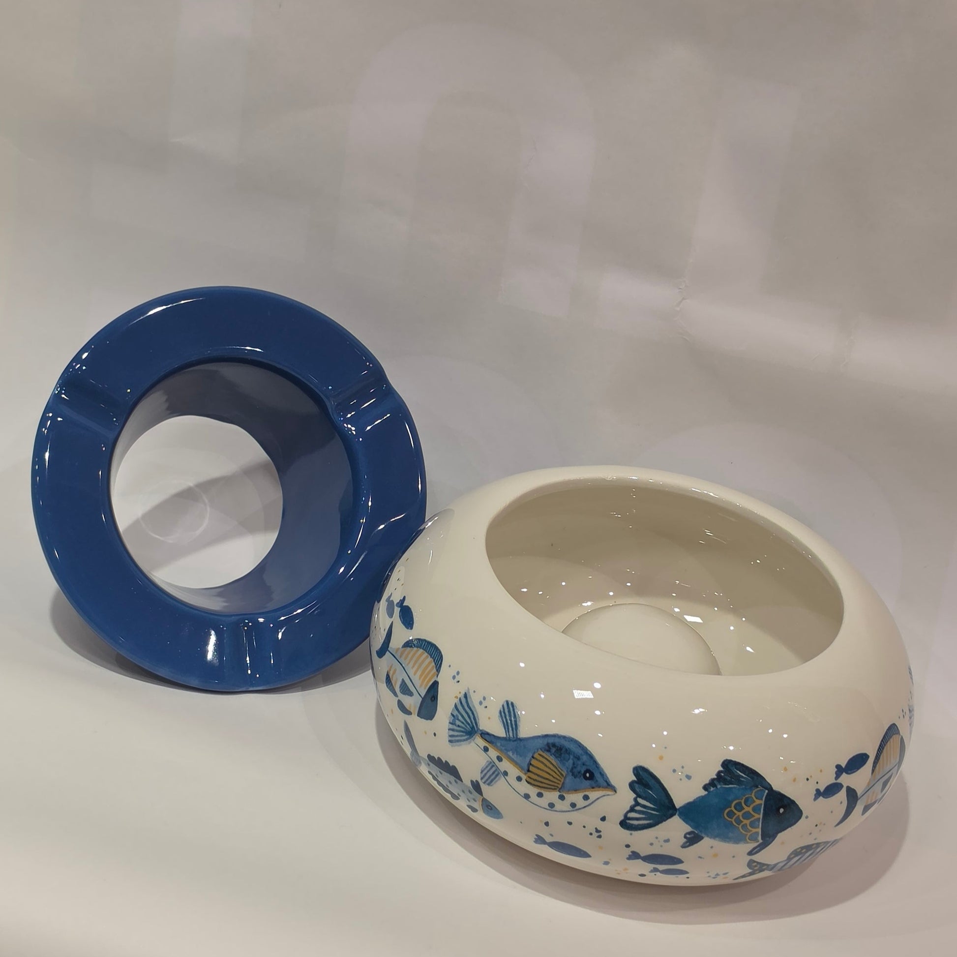 Posacenere antivento in ceramica con coperchio rimovibile blu lucido, decorato esternamente con un motivo artistico di pesciolini dipinti nelle tonalità dell'azzurro e del giallo. La struttura bombata color panna presenta tre scanalature per appoggiare le sigarette, rendendo questo accessorio per fumatori sia funzionale che perfetto come originale complemento d'arredo a tema marino.