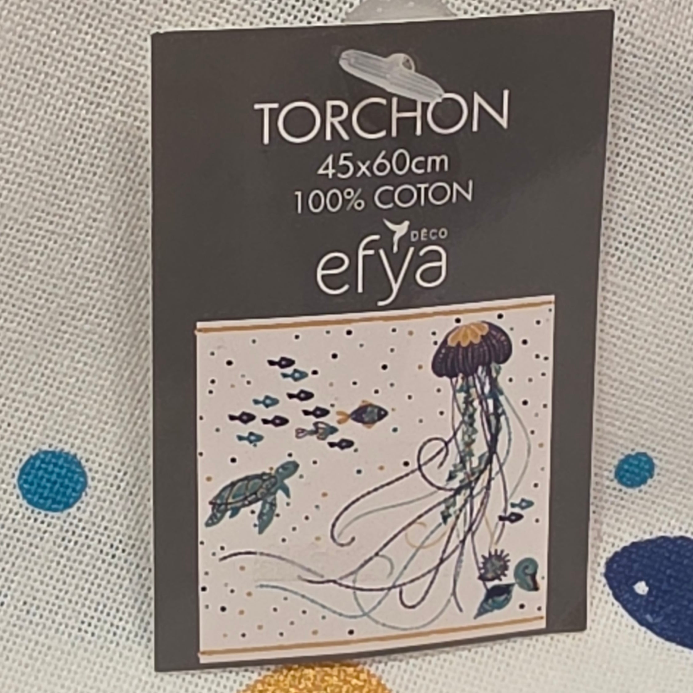 Strofinaccio da cucina in puro cotone del marchio Efya, caratterizzato da una vivace fantasia marina con pesci stilizzati e una medusa stampati nei toni del blu, azzurro e giallo ocra. Il canovaccio presenta un fondo bianco decorato da piccoli pois colorati sparsi, completato da una riga ocra sul bordo inferiore e da una pratica asola scura per appenderlo.