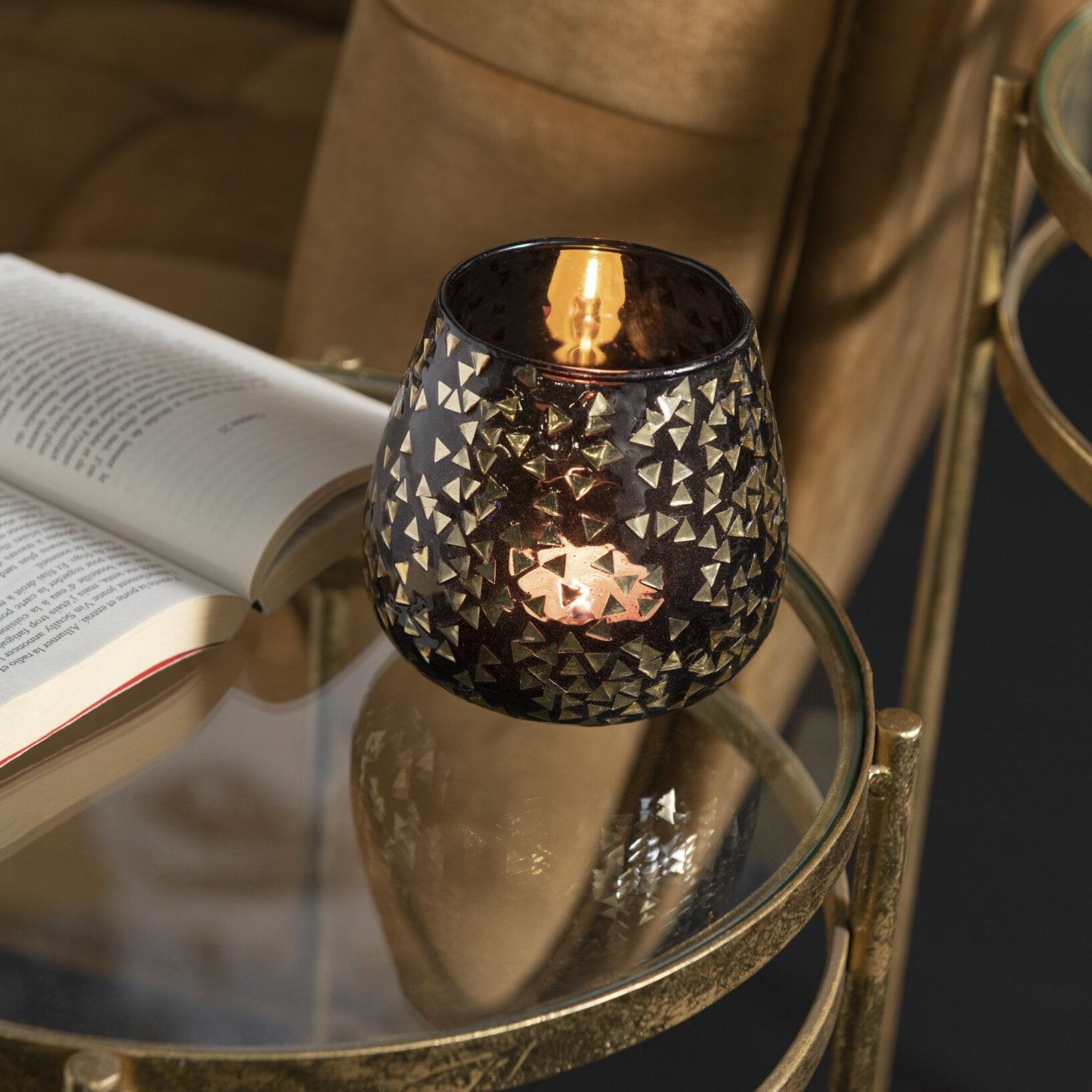 Portacandele decorativo in vetro nero semitrasparente caratterizzato da una raffinata trama geometrica a piccoli triangoli dorati che rivestono l'intera superficie bombata. Questo elegante bicchiere portacandela lascia trasparire una luce calda e soffusa, rendendolo un complemento d'arredo di design ideale per creare un'atmosfera accogliente in casa.
