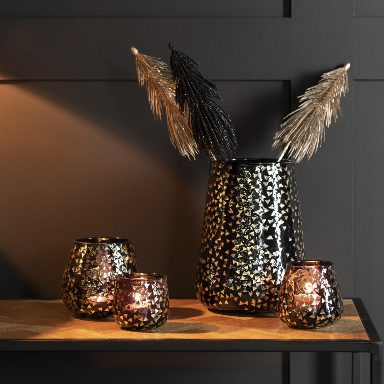 Portacandele decorativo in vetro nero semitrasparente caratterizzato da una raffinata trama geometrica a piccoli triangoli dorati che rivestono l'intera superficie bombata. Questo elegante bicchiere portacandela lascia trasparire una luce calda e soffusa, rendendolo un complemento d'arredo di design ideale per creare un'atmosfera accogliente in casa.