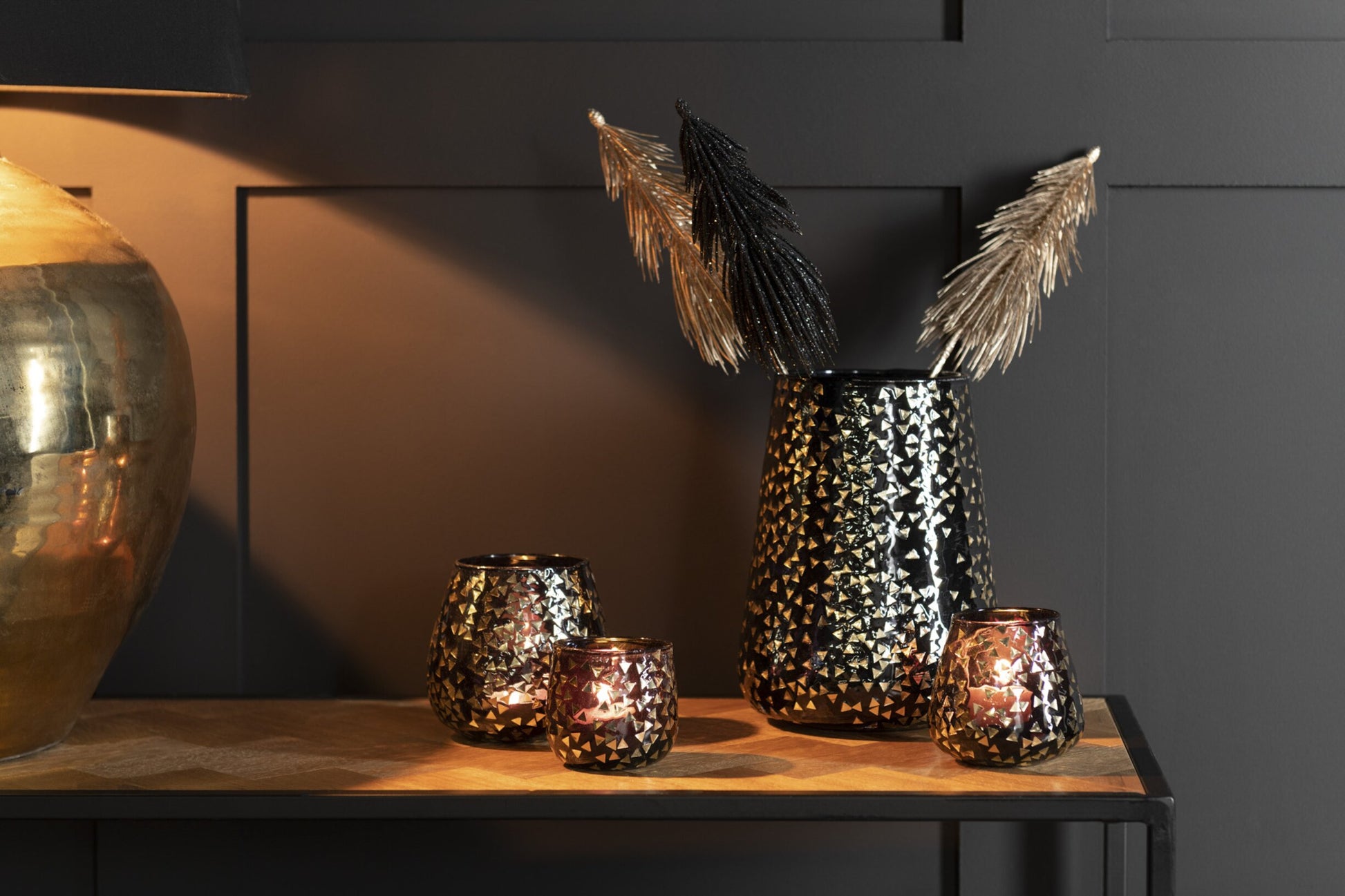Portacandele decorativo in vetro nero semitrasparente caratterizzato da una raffinata trama geometrica a piccoli triangoli dorati che rivestono l'intera superficie bombata. Questo elegante bicchiere portacandela lascia trasparire una luce calda e soffusa, rendendolo un complemento d'arredo di design ideale per creare un'atmosfera accogliente in casa.