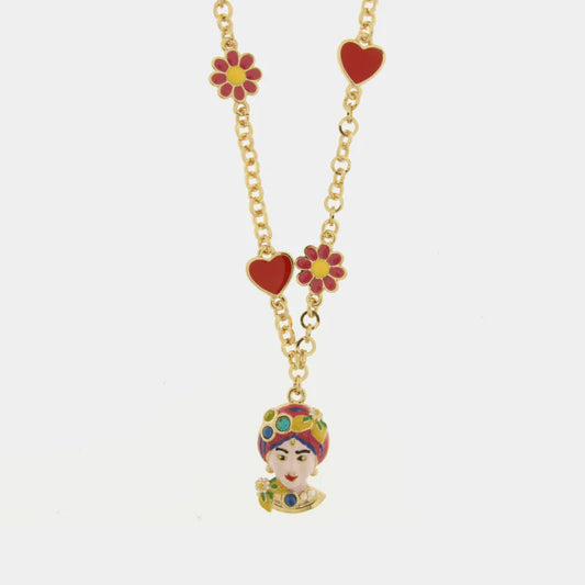 Questa vivace collana dorata della collezione Etna si distingue per il raffinato pendente a forma di Testa di Moro, decorato a mano con smalti multicolore e dettagli floreali. La catena a maglie tonde è arricchita da charms smaltati a forma di cuori rossi e fiori rosa con centro giallo, che donano un tocco romantico e solare al gioiello. Perfetta per chi ama lo stile folk-chic, questa collana con Testa di Moro rosa celebra la tradizione siciliana con un design moderno e accattivante firmato bysimon.