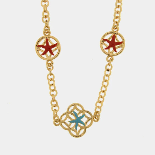 Collana lunga in metallo con finitura dorata del marchio BySimon appartenente alla collezione Fiori di Lava caratterizzata da una catena a maglia rolo impreziosita da tre elementi traforati ispirati alle maioliche mediterranee. Il gioiello presenta un modulo centrale a forma di quadrifoglio che ospita una stella marina smaltata in azzurro, affiancato lungo la catena da due charms circolari con stelle marine smaltate in un vivace colore rosso. 