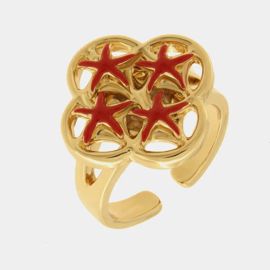 Anello regolabile con finitura dorata del marchio BySimon appartenente alla collezione Fiori di Lava caratterizzato da un castone superiore a forma di quadrifoglio con lavorazione traforata ispirata alle maioliche mediterranee. Il design è decorato da quattro stelle marine smaltate in un vivace colore rosso, posizionate simmetricamente all'interno dei petali della struttura. La fascia dell'anello è lucida e presenta un gambo aperto sul retro per consentire la regolazione della misura, rendendo questo gioiel